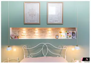 Cozy bedroom with teal walls, a white metal bed, two pendant lamps, and a glowing display shelf above the headboard containing books and a clock.